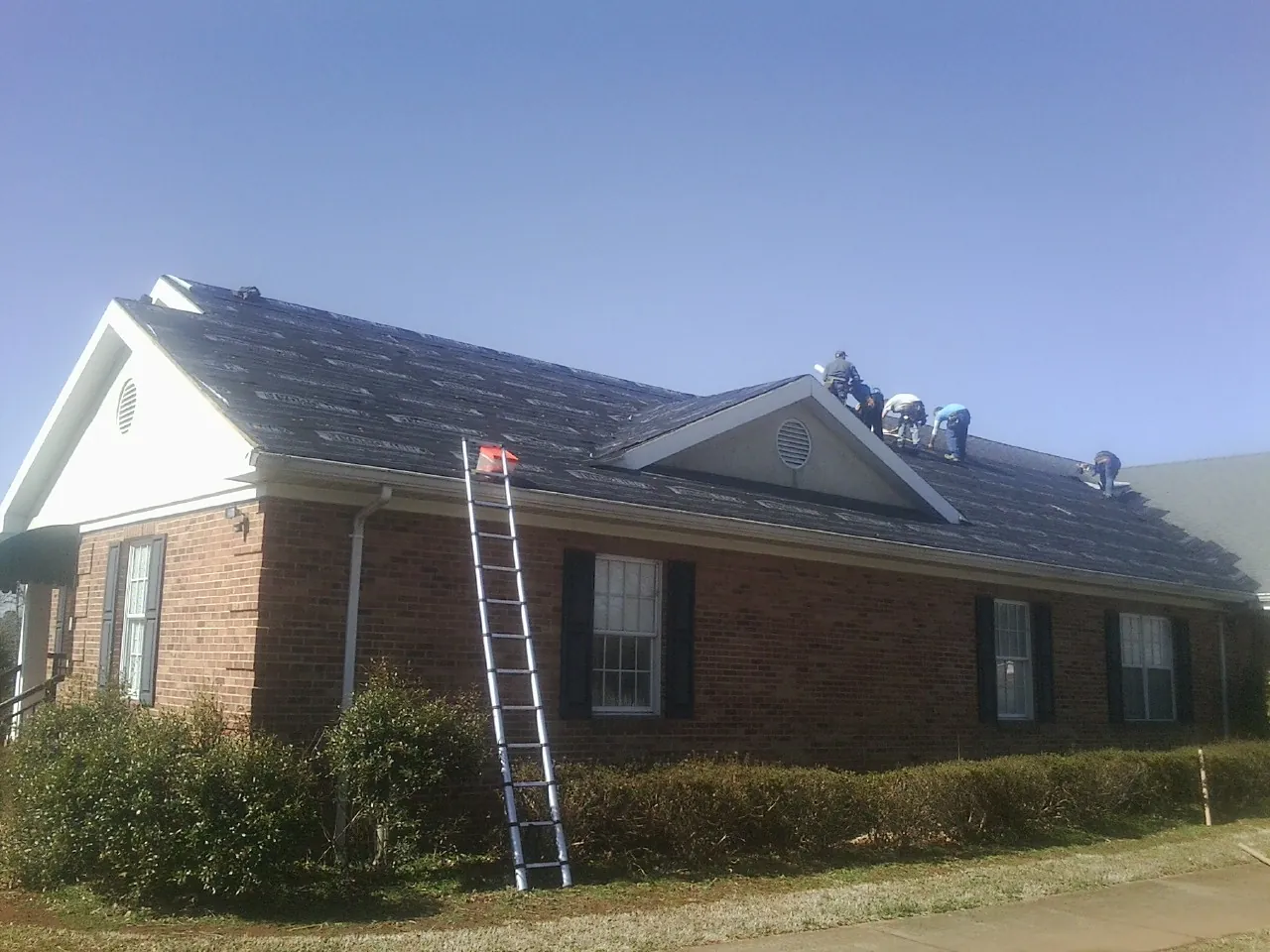 Crew tearing shingles off a brick ranch roof with a ladder set — Emergency Roof Repair work by our crew in Bay Shore