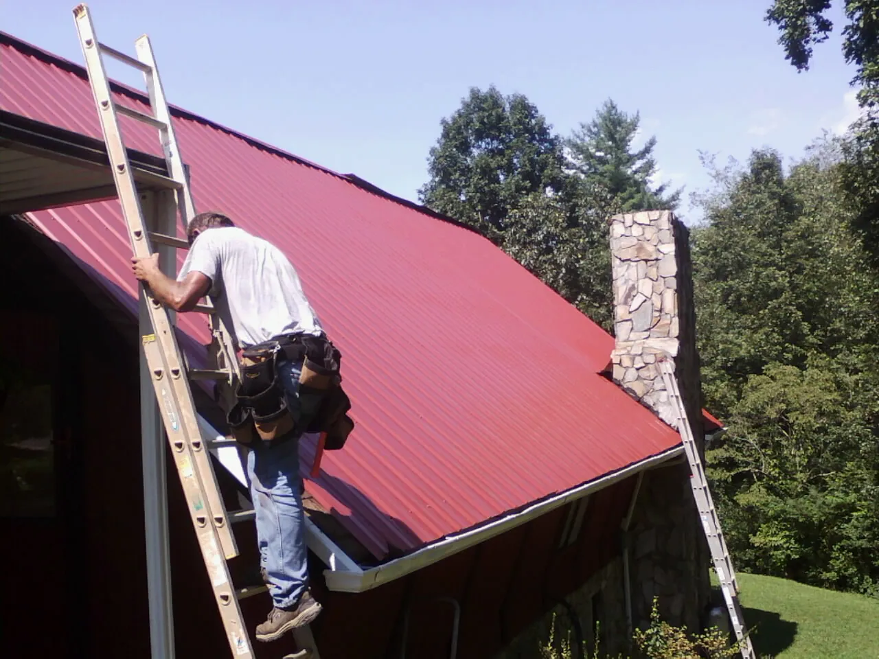 Roofer on a ladder beside a red metal roof and stone chimney — Soffit & Fascia Repair by Roofing Brothers in Bay Shore